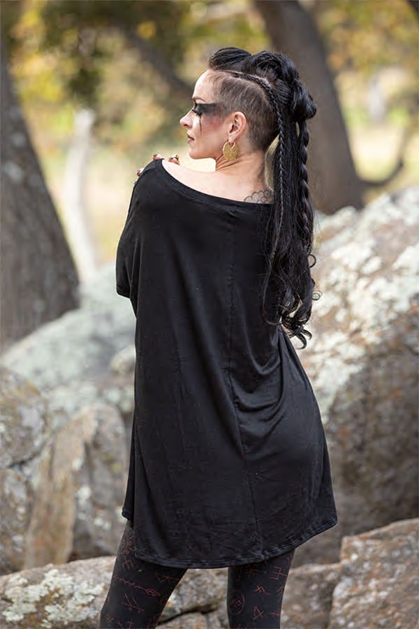 A person with long black braided hair and shaved sides models the oversized black Luna Long Sleeve Runes shirt by Melodia Designs and patterned leggings while standing on rocks outdoors, facing away from the camera.