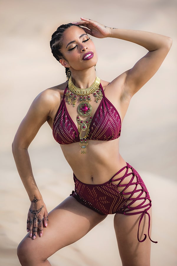 A woman poses on the sand in the Melodia Designs Lexi Luxor Halter Bra, a maroon patterned triangle bikini top with high-waisted lace-up sides, accessorized with bold gold jewelry and a statement necklace, her eyes closed and hand on forehead.