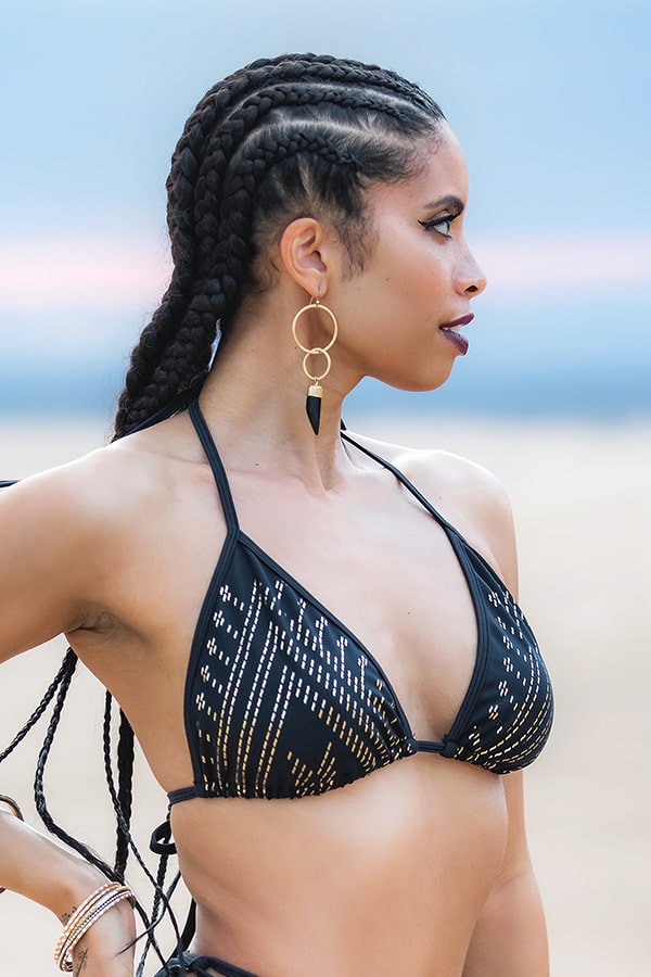 A woman with long braided hair wears the Melodia Designs Lexi Luxor Halter Bra in black and gold shimmer, paired with large hoop earrings, looking to the side on a beach with a blurred sky and sand background.