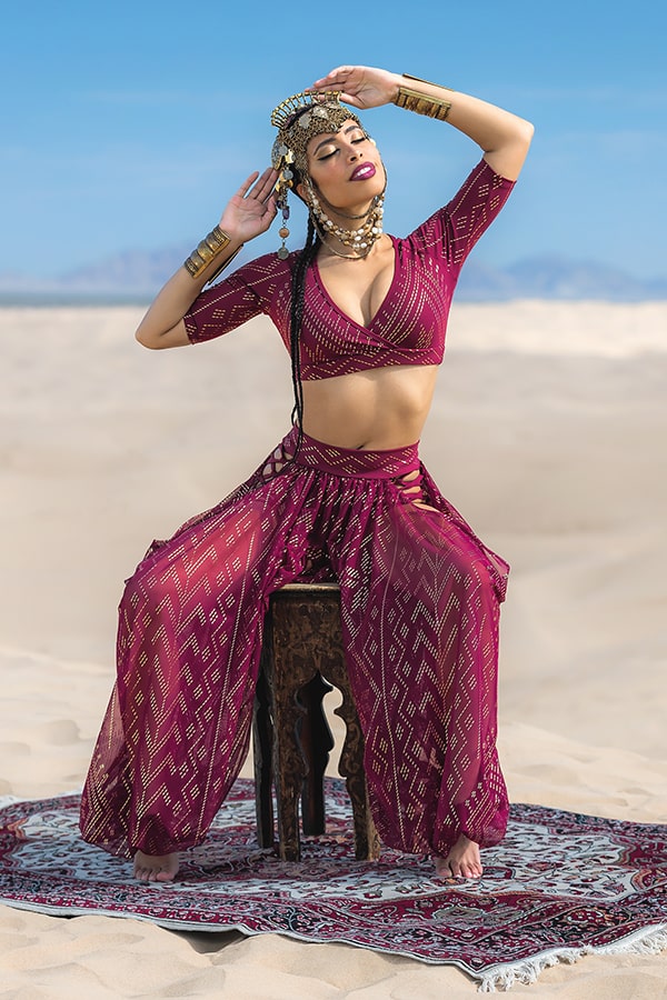 A woman wearing Melodia Designs’ Sahara Peekaboo Luxor Pantaloon poses on a wooden stool atop a patterned rug in the desert, adorned with ornate jewelry and a headpiece, arms raised and eyes closed beneath the clear blue sky.