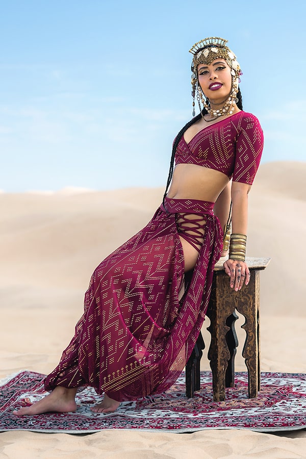 A woman in Melodia Designs’ Sahara Peekaboo Luxor Pantaloon, featuring a burgundy and gold pattern and sheer hip drape, accessorized with gold jewelry and headdress, sits on a stool atop a patterned rug in the desert under a blue sky.