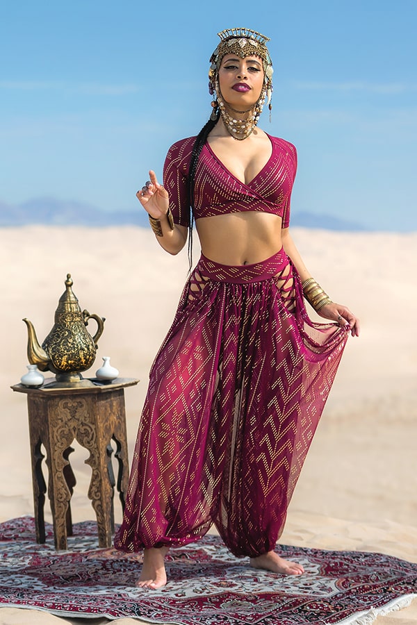 A woman in a jeweled headdress and Melodia Designs’ Sahara Peekaboo Luxor Pantaloon with an elastic-free waistband poses in the desert on a rug beside a wooden table with an ornate teapot, framed by blue sky and sand dunes.