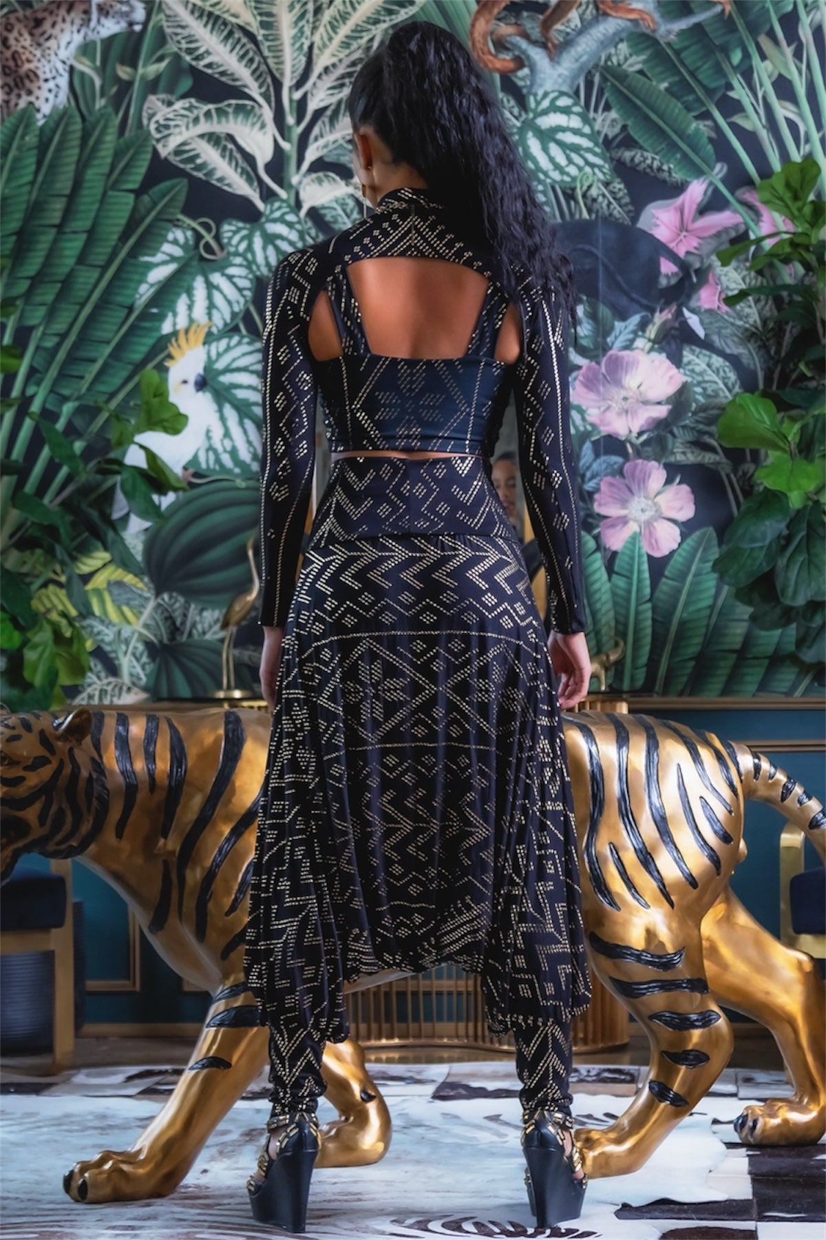 Wearing the Christine Luxor Harem Jumper by Melodia Designs, a person stands with their back to the camera before a gold tiger statue, surrounded by lush green plants and a floral mural.