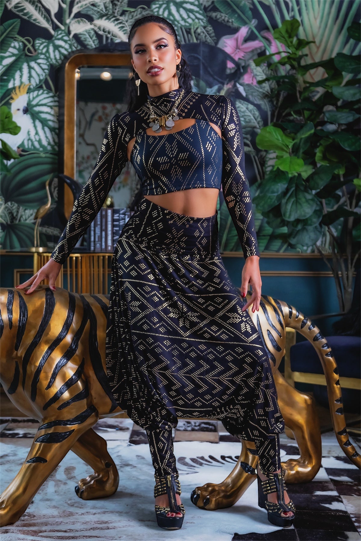 A woman poses confidently indoors in the Christine Luxor Harem Jumper by Melodia Designs, leaning against a large gold tiger statue with lush green plants, patterned wallpaper, and a decorative mirror in the background.