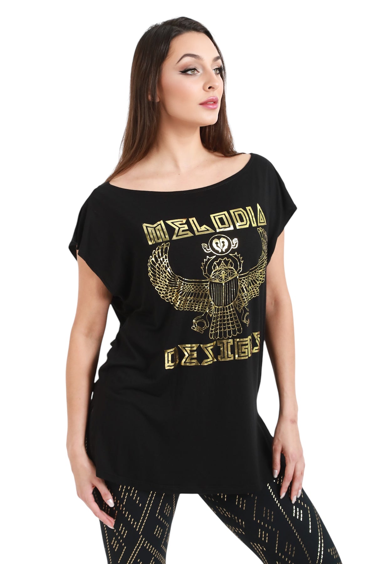A woman models the Melodia Designs Scarab Tunic, a loose black top with gold Egyptian scarab motifs and branding, paired with eco-luxe Modal black leggings featuring gold geometric patterns, against a white background.