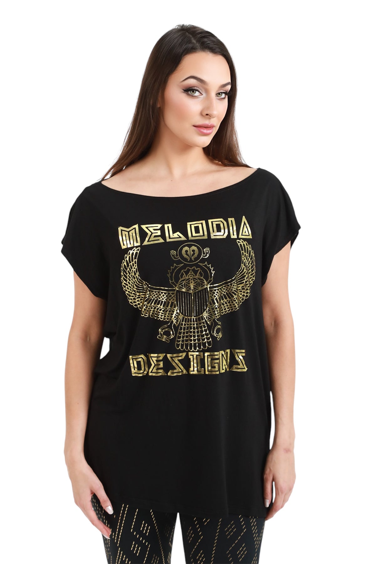 A woman with long brown hair stands facing forward against a white background, wearing the Scarab Tunic by Melodia Designs—a loose black eco-luxe Modal T-shirt featuring gold Melodia Designs lettering and an Egyptian winged scarab design on the front.