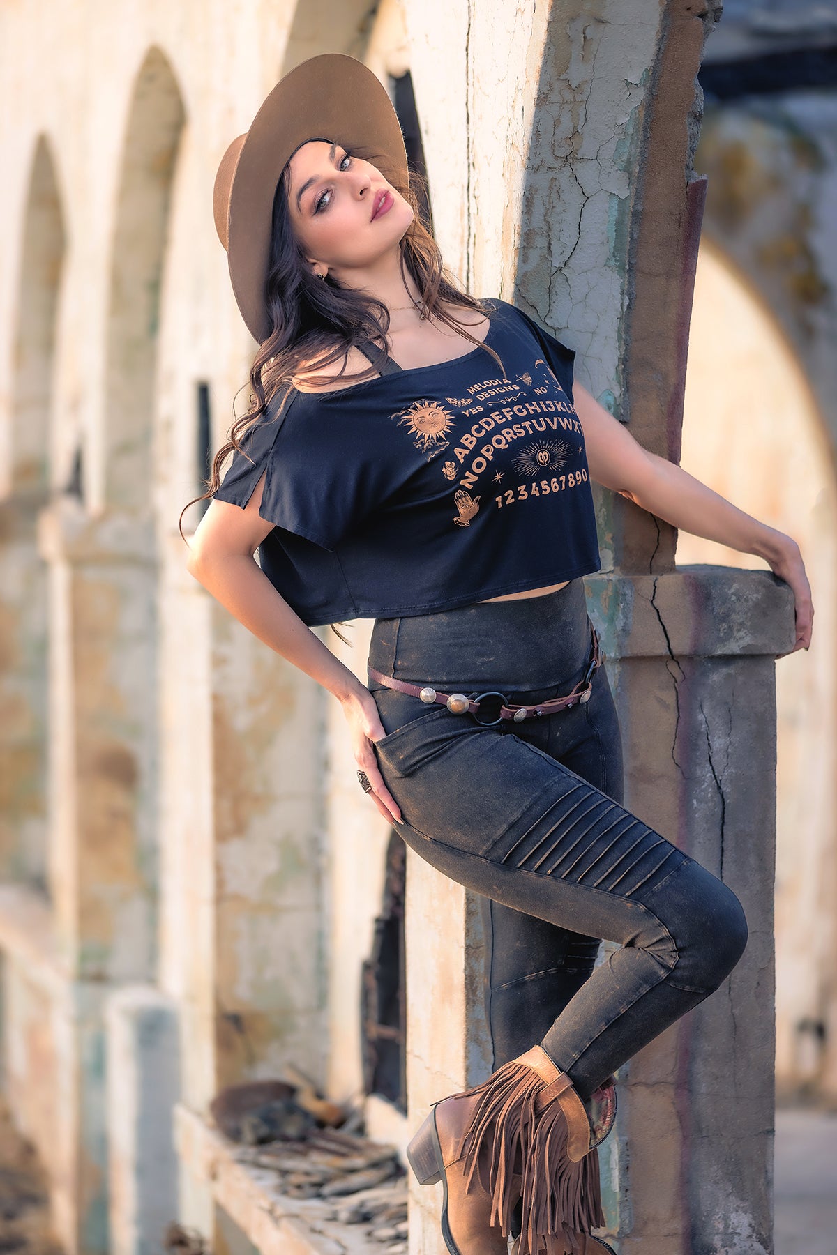 A woman wearing the Melodia Designs Ouija Dede Crop and a wide-brimmed hat leans against a weathered stone archway outdoors, one leg bent and head tilted back as soft sunlight highlights her fringed boots and relaxed pose.