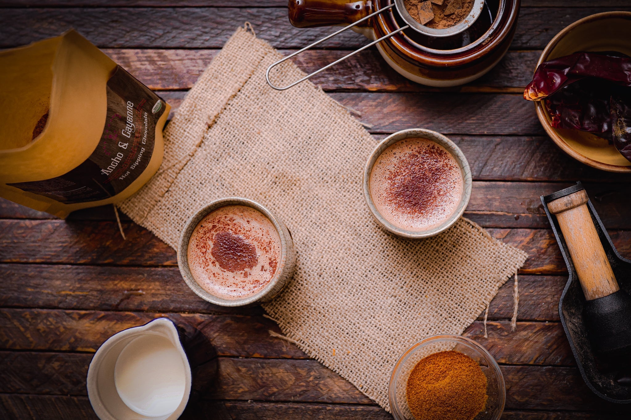 Two mugs of Ancho & Cayenne Sipping Chocolate from chocolita.com, topped with cocoa powder, rest on burlap with a jug of milk, brown sugar, spices, and a cocoa powder bag—ideal for enjoying an authentic Mayan-inspired treat.