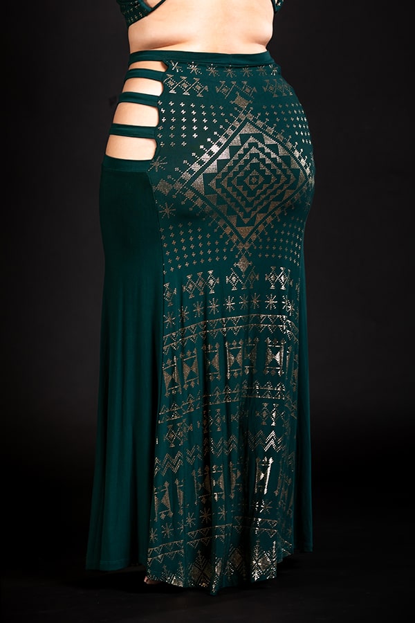 A person wears the Alkimiya Persephone Skirt Malachite by Melodia Designs—an eco-luxe, long dark green skirt with metallic gold geometric print and side cut-outs—standing against a black background.