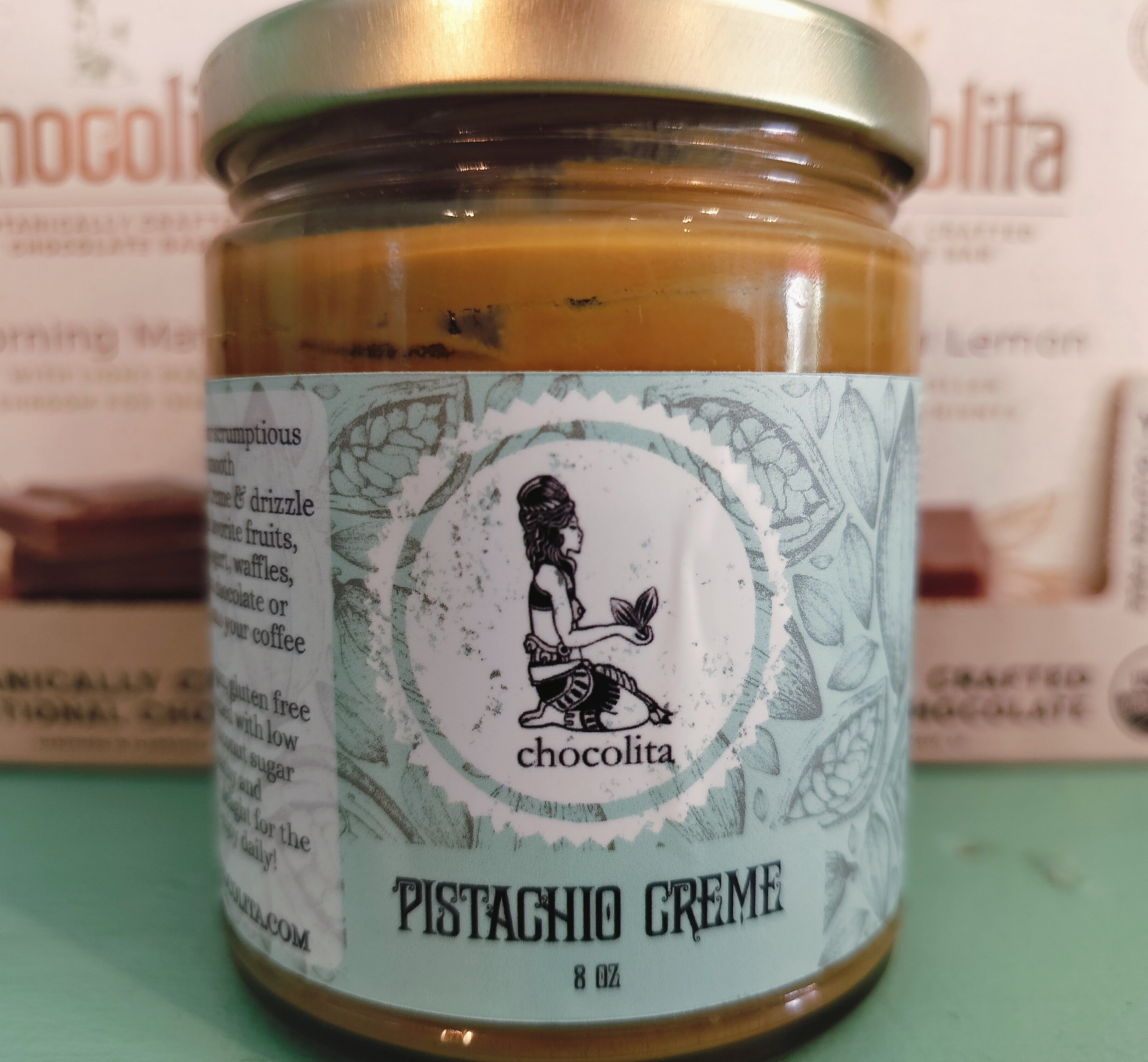 An 8 oz glass jar of chocolita.com Pistachio Creme with a green lid. The label shows a cross-legged woman among leaves. This creamy spread is made from stone ground pistachios.