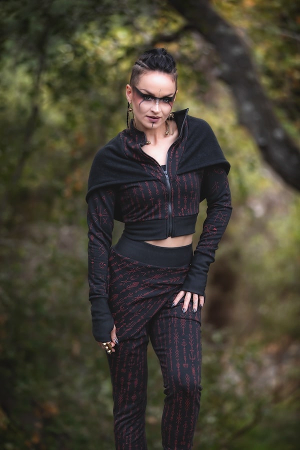 Wearing dark makeup and black lipstick, a person stands in a forest dressed in Melodia Designs Freydis Hoodie Runes—an eco-luxe black and red patterned outfit—surrounded by trees and lush greenery.