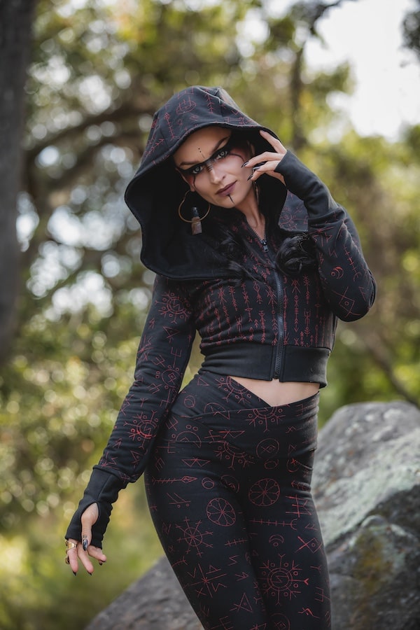 Wearing the Freydis Hoodie Runes by Melodia Designs, this person poses outdoors among trees and rocks, showcasing dramatic makeup and jewelry for a mystical, alternative vibe.