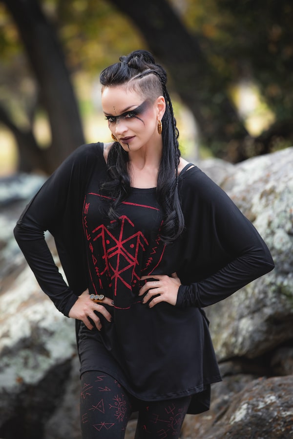 A woman with long black hair, side shaved, wearing dramatic black eye makeup and a Melodia Designs Luna Long Sleeve Runes with red bindrune-inspired design, stands outdoors among rocks and trees with her hands on her hips.