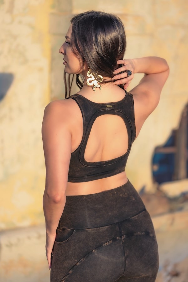 A woman with long dark hair stands outdoors, facing away with one hand on her head, wearing a sleeveless black Rustique Serena Bra 2.0 by Melodia Designs and matching leggings, accessorized with large earrings and a ring.