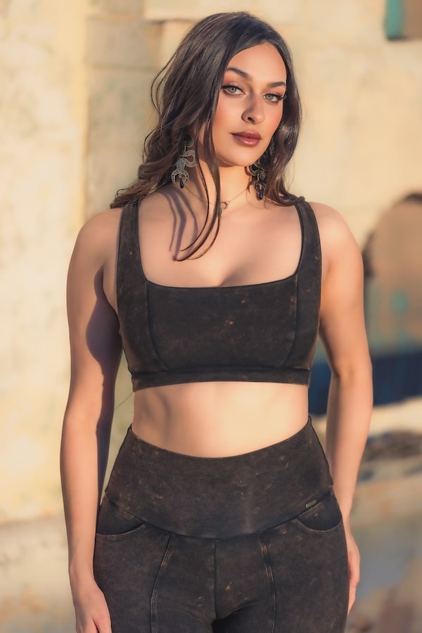 A woman with long brown hair wears the Melodia Designs Rustique Serena Bra 2.0 crop top and matching high-waisted pants outdoors. She pairs statement earrings and natural makeup, exuding confidence and style in eco-luxe fabric.