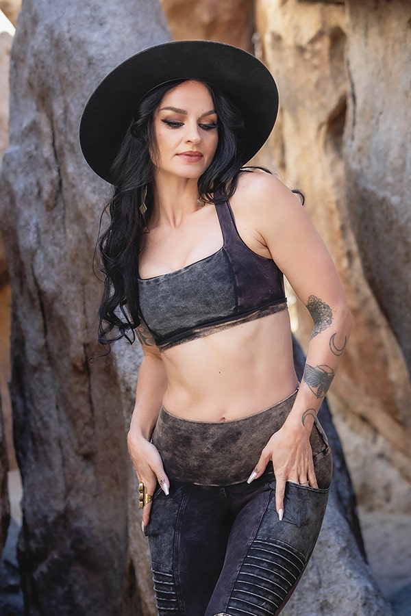 A woman with long black hair and tattoos wears the Melodia Designs Serena Bra 2.0 Grunge, styled with a black wide-brimmed hat, dark crop top, and matching leggings. She stands among large rocks, looking down with one hand on her thigh.