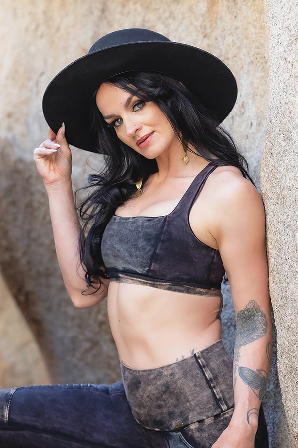 A woman with long dark hair and tattoos on her left forearm poses confidently against a rock wall, wearing Melodia Designs’ Serena Bra 2.0 Grunge in eco-luxe fabric, styled with a black wide-brimmed hat, crop top, and matching pants.