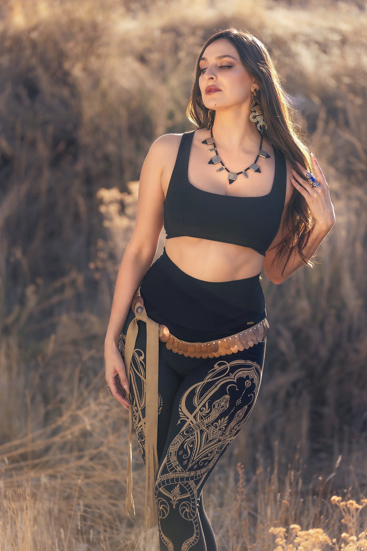 A woman stands in tall dry grass, sunlight on her hair, wearing Melodia Designs Elsa Legging Serpent with a high-rise waistband and a black crop top, accessorized with a necklace, earrings, and a coin belt.