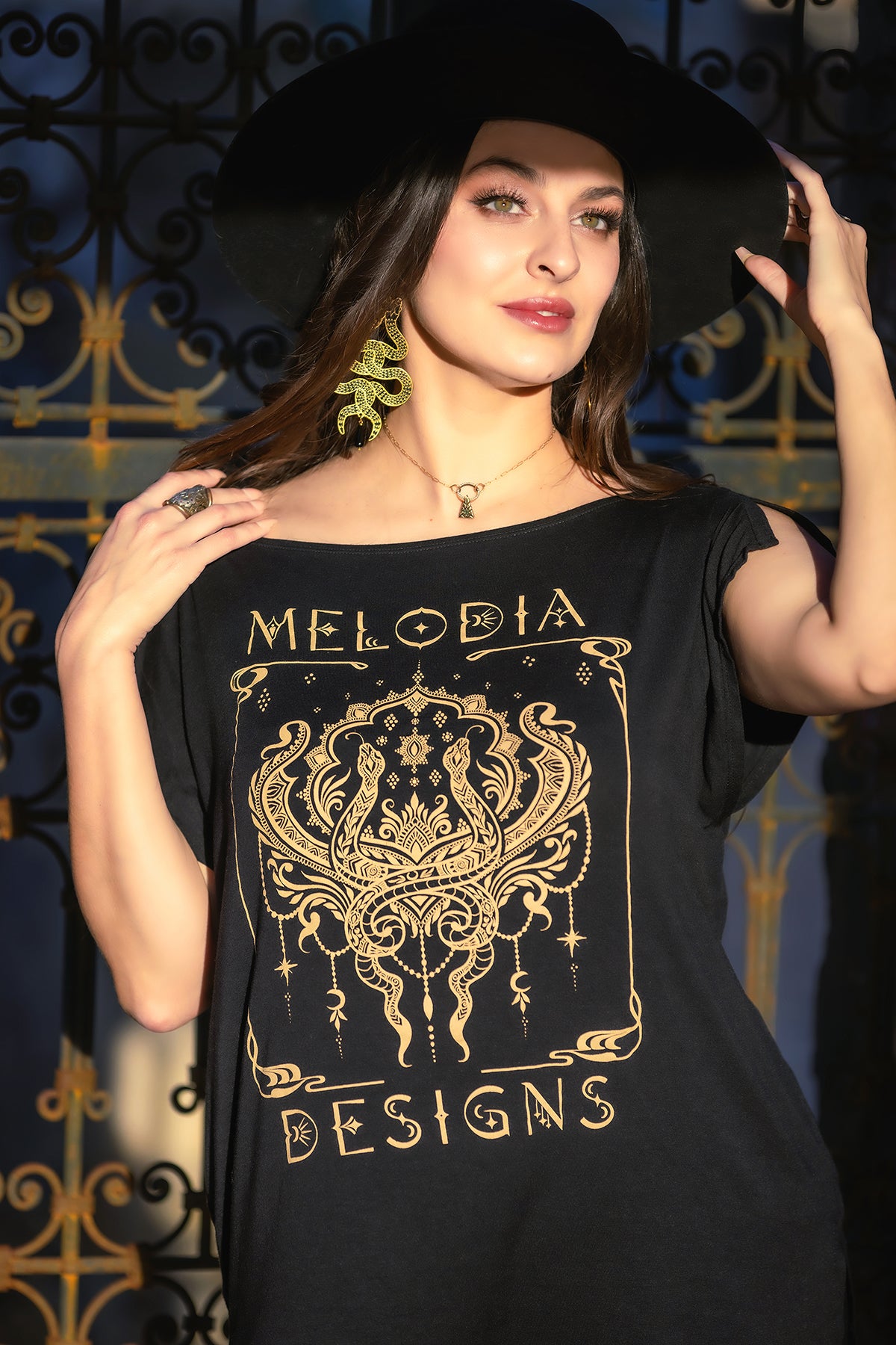 A woman in the Melodia Designs Nouveau Serpent Tunic, a black eco-luxe T-shirt with a gold ornate logo, paired with a wide-brim hat, poses by a decorative iron gate as sunlight highlights her face and earrings.