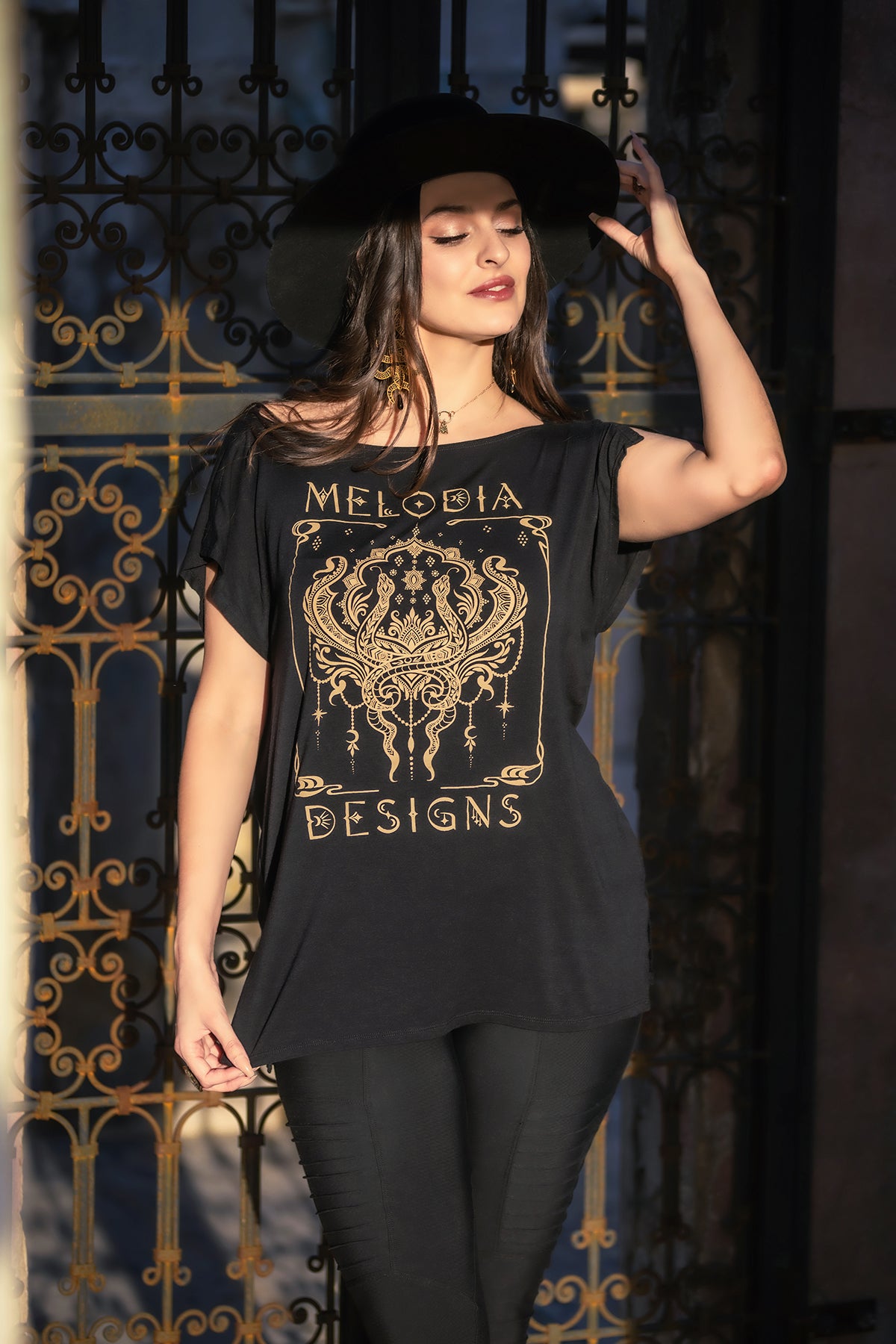 A woman in front of ornate iron gates wears a black wide-brimmed hat and the Melodia Designs Nouveau Serpent Tunic, made from eco-luxe fabric. She holds her hat with one hand and gazes slightly to the side.