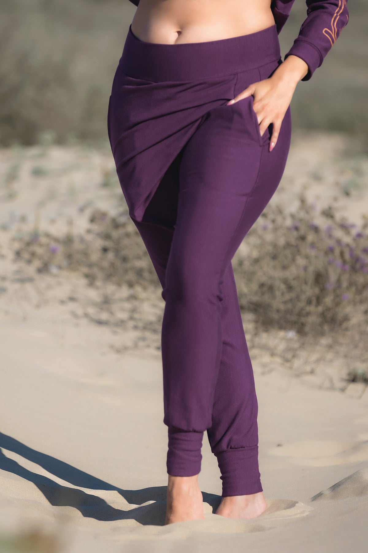 A barefoot person stands on sand, wearing Melodia Designs' Shiva Wrap Jogger in Aubergine, made from eco-friendly bamboo with a high waist and wrap detail. Their hand is in one pocket; only their lower half is visible.