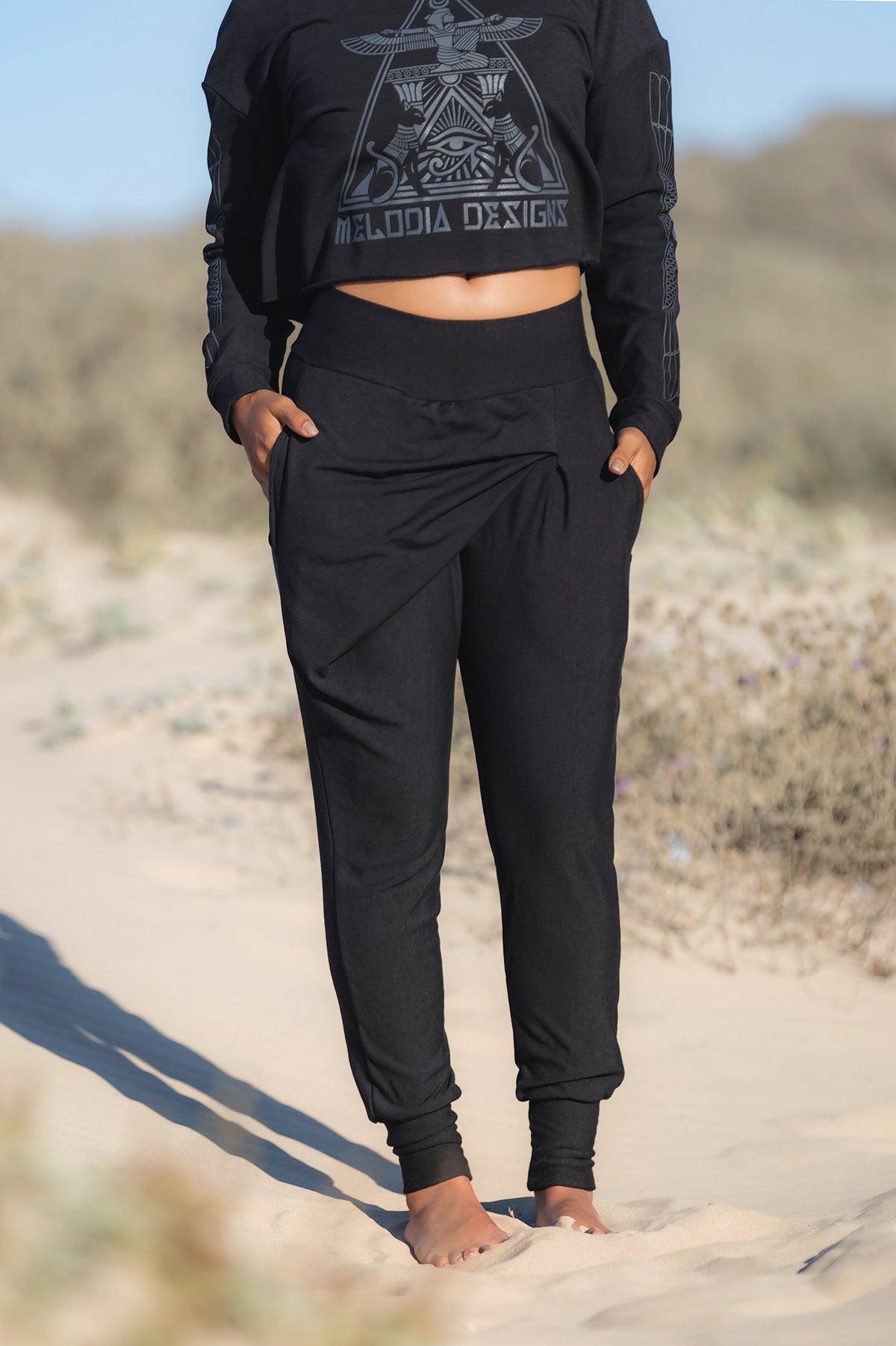 A person stands on a sandy path in Melodia Designs' Shiva Wrap Jogger Black pants and a black geometric long-sleeve crop top, enjoying eco-friendly comfort amid desert vegetation with hands in their pockets.