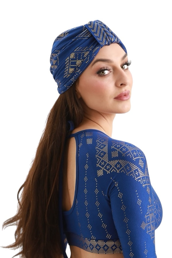 A woman with long brown hair wears a royal blue Melodia Designs Urban Turban with a loop front for versatile styling and a matching gold-printed top, glancing over her shoulder against a white background.