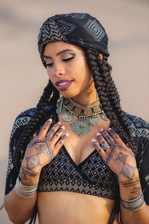 A woman with long braids wears the versatile Urban Turban by Melodia Designs, traditional jewelry, and henna on her hands. In a black patterned top, she gazes down serenely.
