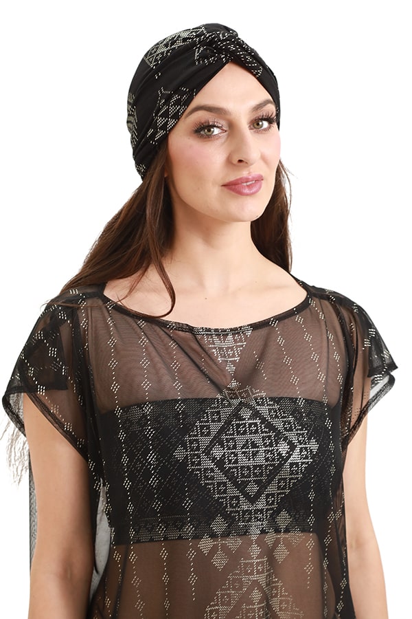 A woman wearing the Urban Turban by Melodia Designs—a versatile black and silver headwrap—paired with a sheer black top with silver geometric designs, poses and smiles against a white background for a chic, minimalist look.
