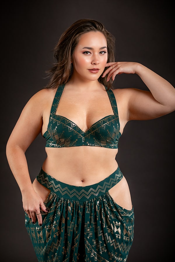 A woman with medium-length dark hair models the Melodia Designs Alkimiya Vixen Bra Malachite—a structured green and gold underwire bra—posing confidently against a dark background.