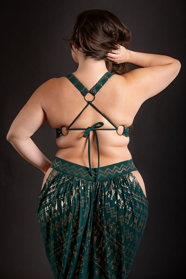A woman stands with her back to the camera, wearing the Melodia Designs Alkimiya Vixen Bra Malachite, featuring green and gold geometric patterns, an open-back design with crisscrossed straps, against a dark background. Her left hand rests on her head.